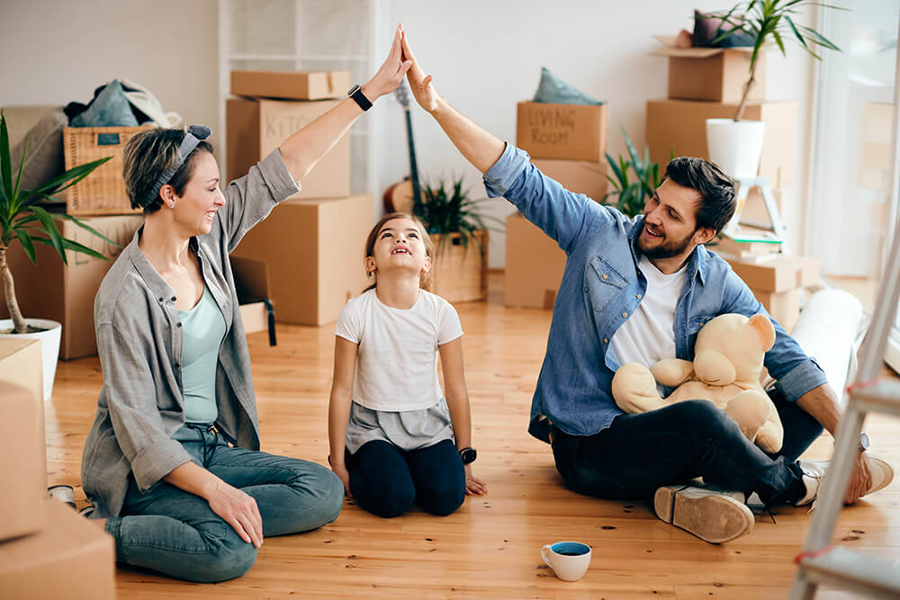 A family sitting on the floor of their new home with a large amount of boxes surrounding them, the parents are high-fiving and their child looks up at their high five.